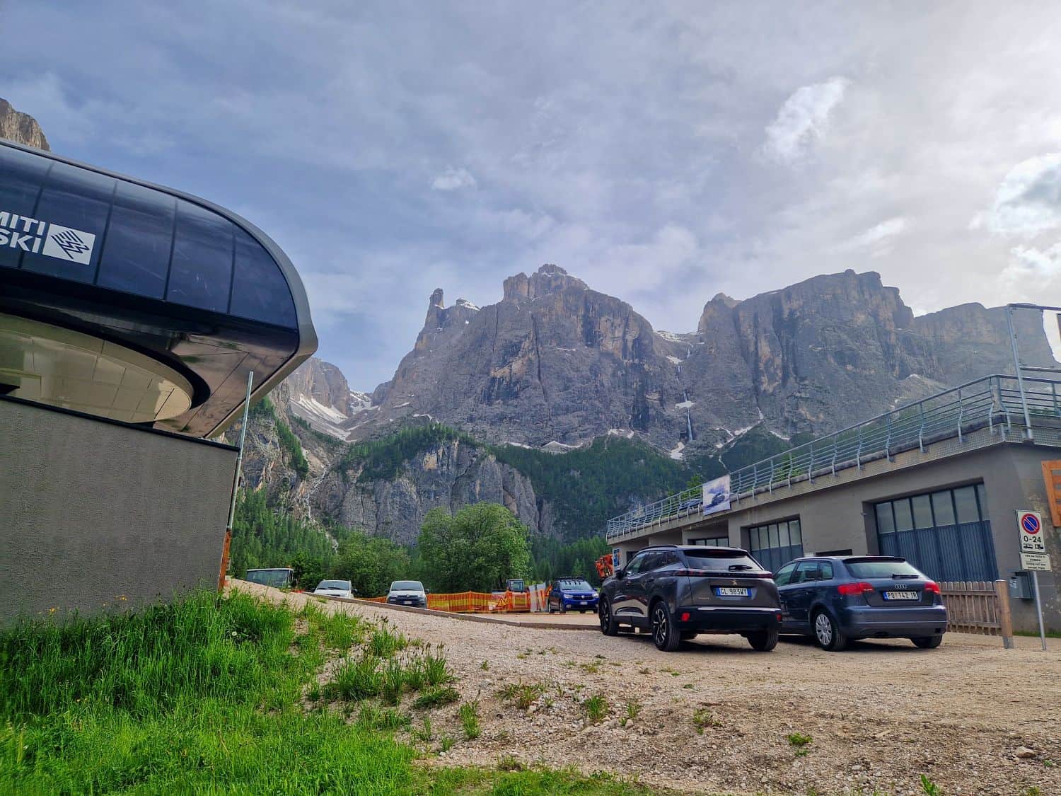 Cars parked near a modern ski lift station, with dramatic rocky mountains and green forests in the background under a partly cloudy sky.