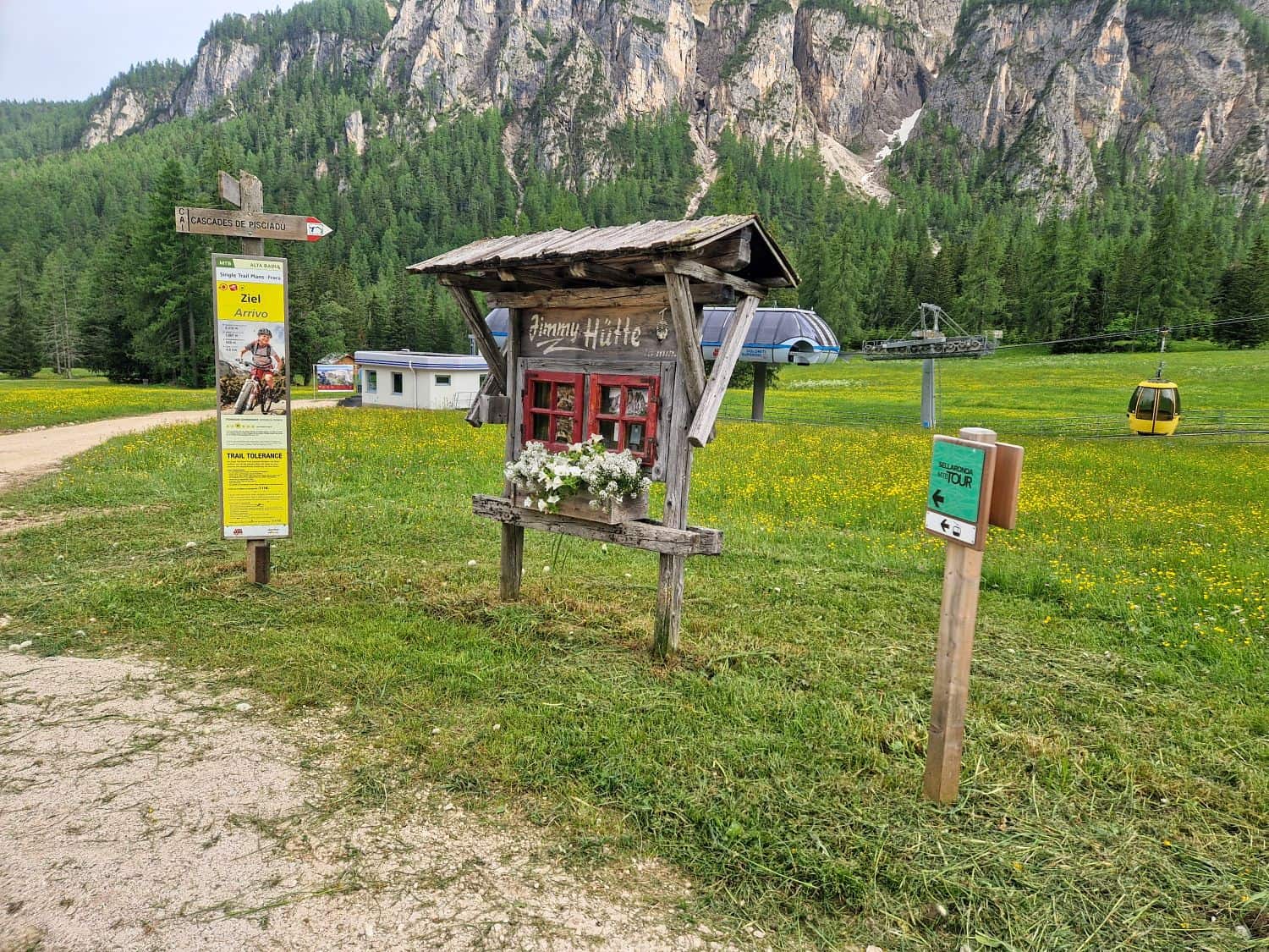 A rustic wooden sign with flowers stands in a grassy field near a yellow info board, a green post, and a gondola; forested mountains and a ski lift are visible in the background.