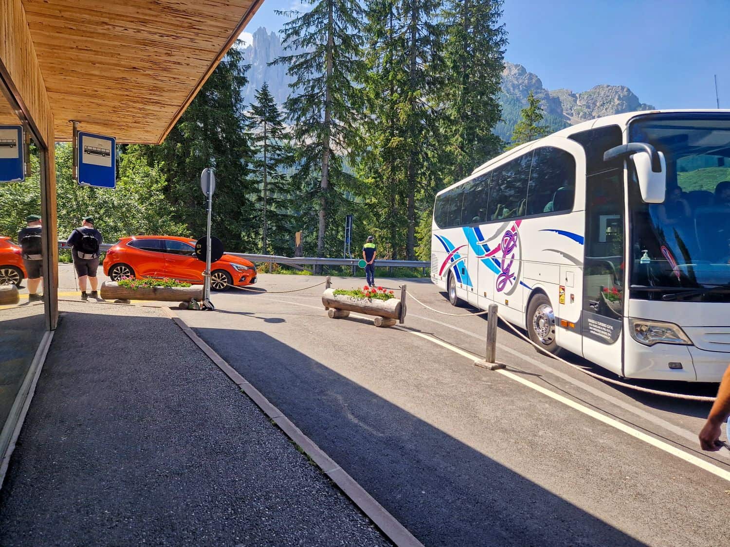 A white tour bus and two orange cars are parked near a wooden building in a mountain area with tall pine trees. A person stands in the middle of the road under a clear, sunny sky.