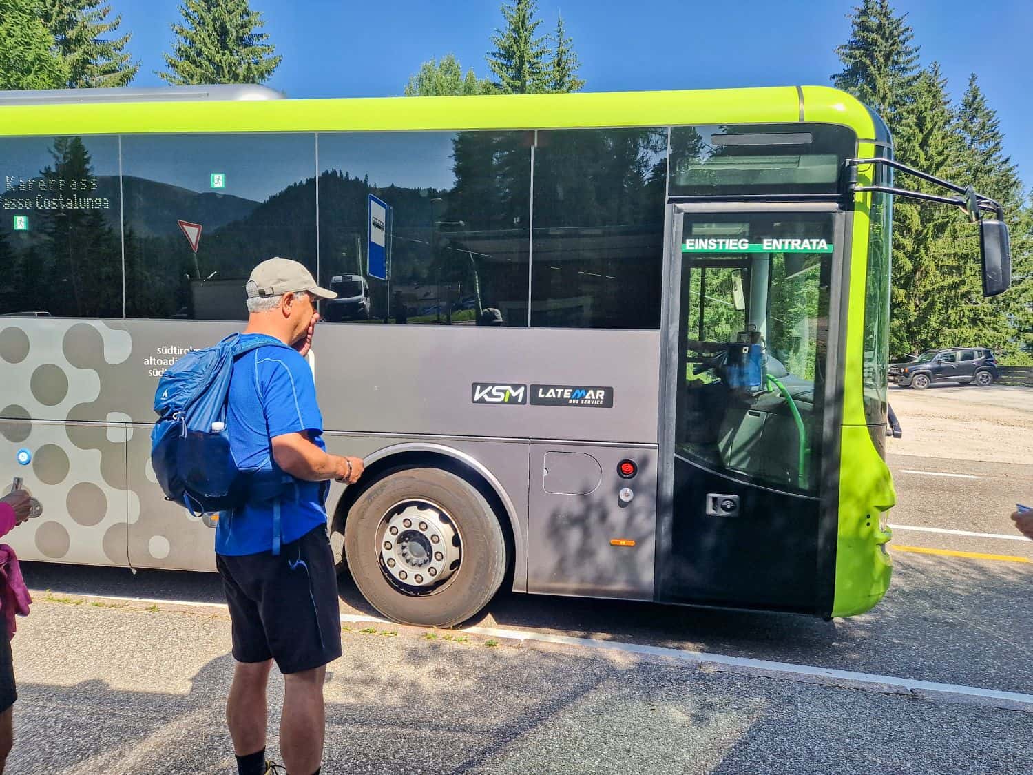 A man wearing a blue shirt and backpack waits beside a modern green and black bus on a sunny day, with trees and mountains visible in the background.