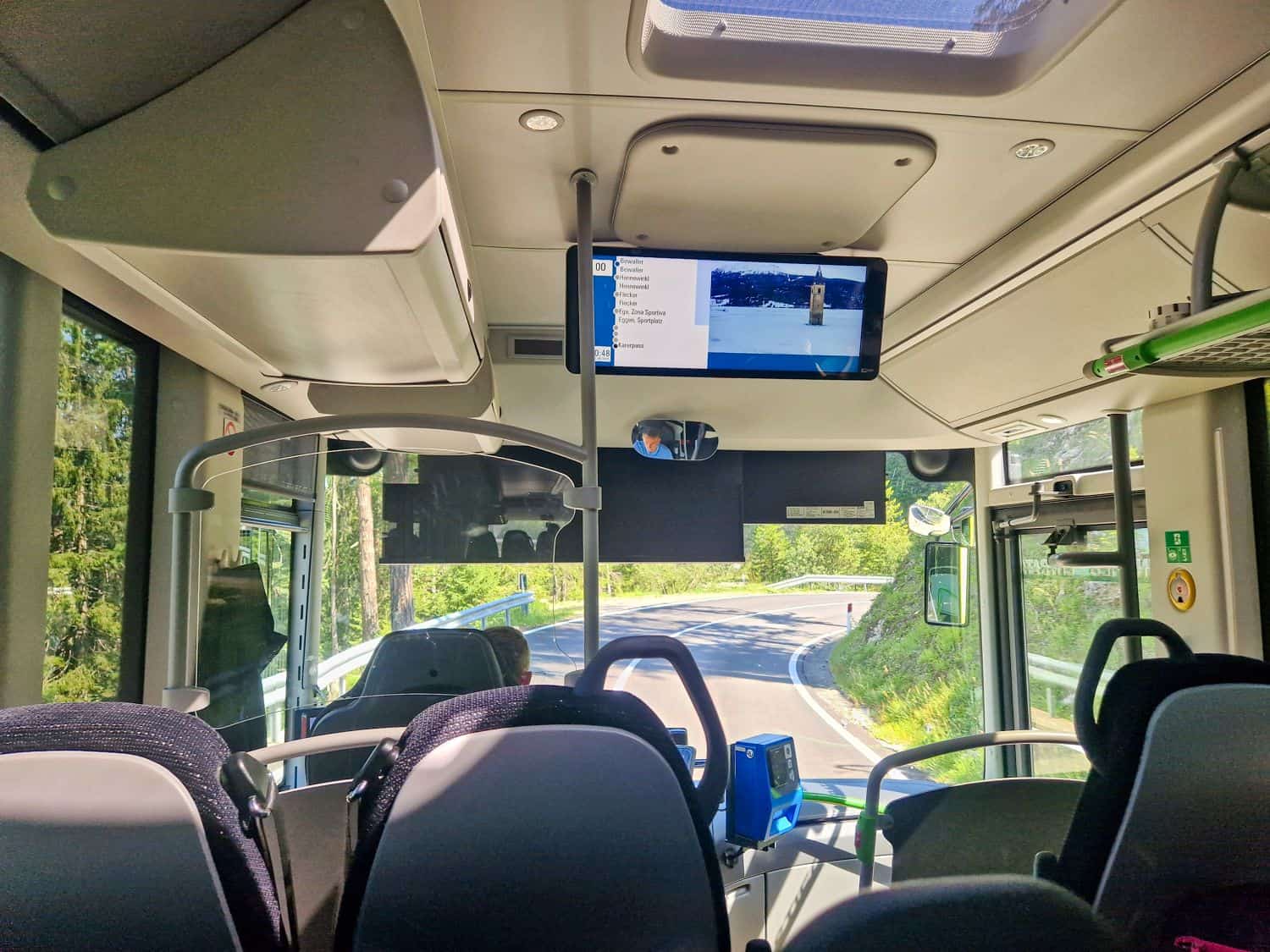Interior of a modern bus with empty seats, a screen showing information above the windshield, and a curving road with greenery visible through the front and side windows. Sunlight streams in from the windows and skylight.