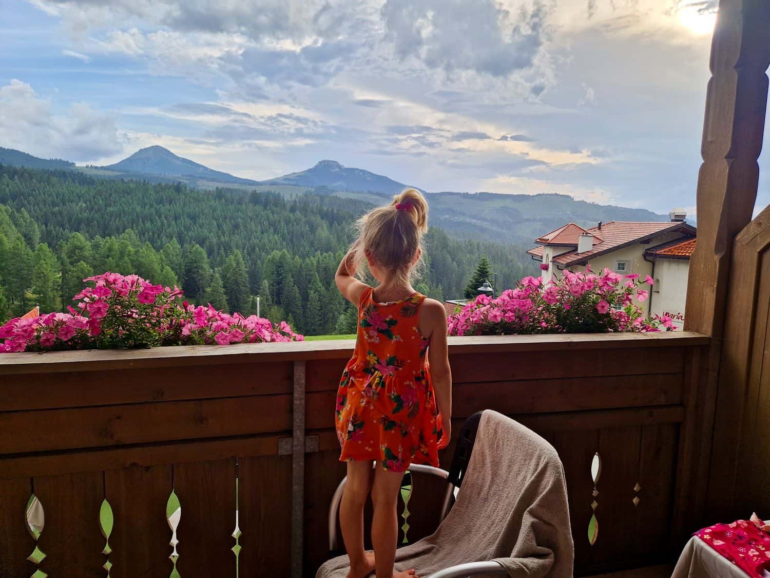 A young girl in an orange dress stands on a chair on a balcony, looking out at a scenic view of mountains, pine forests, and a cloudy sky. Pink flowers line the balcony railing.