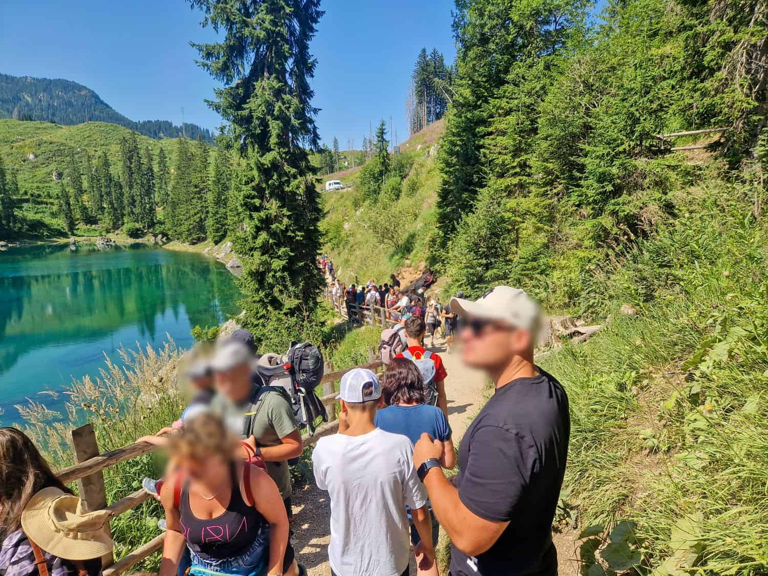 A group of people, some with backpacks and hats, walk along a narrow dirt trail beside a clear lake surrounded by green hills and tall trees under a bright blue sky.