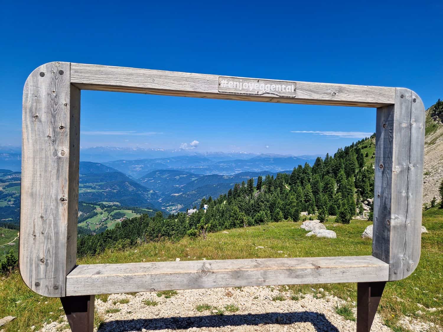A large wooden frame labeled “#enjoyeggental” stands on a grassy mountain slope, perfectly framing a scenic view of green hills, forests, and distant blue mountains under a clear sky.