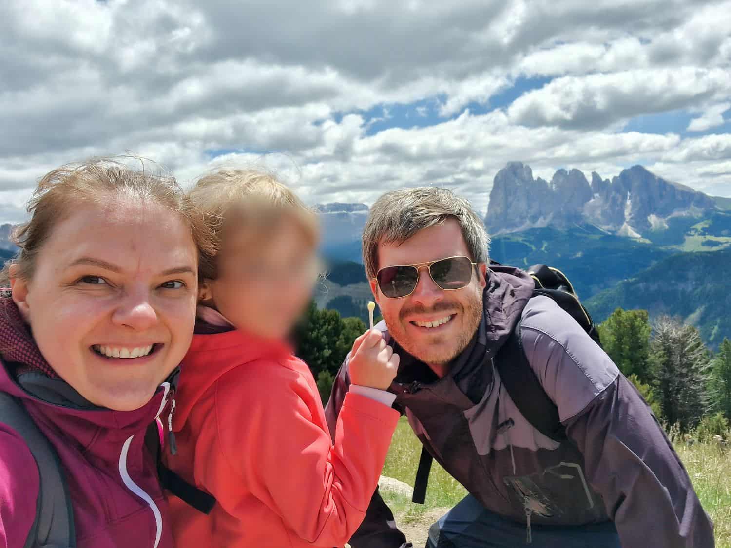 A smiling woman, a man in sunglasses, and a young child in outdoor jackets pose for a selfie on a grassy hillside with mountains and a partly cloudy sky in the background. The child's face is blurred.