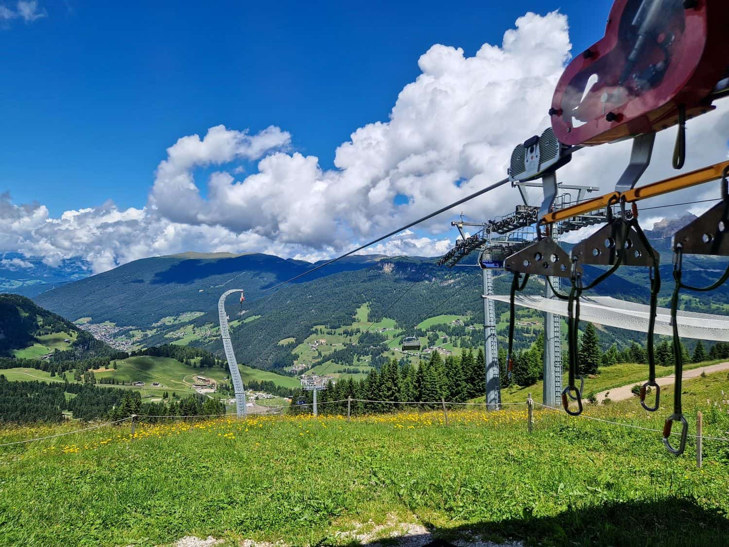 A scenic view of green hills and valleys with a mountain village below, under a blue sky with clouds. In the foreground, a zipline launch platform and equipment are visible.