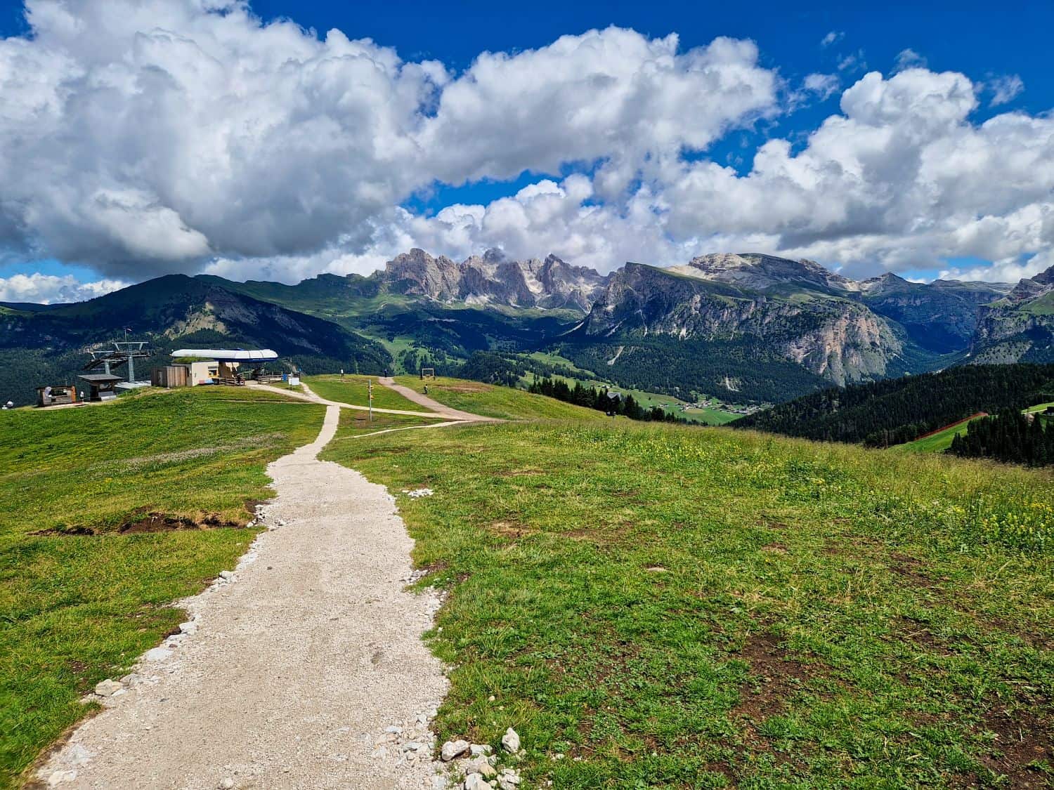 A winding dirt path leads over green hills toward distant rocky mountains under a bright blue sky filled with puffy white clouds. A small building sits on the left side of the landscape.
