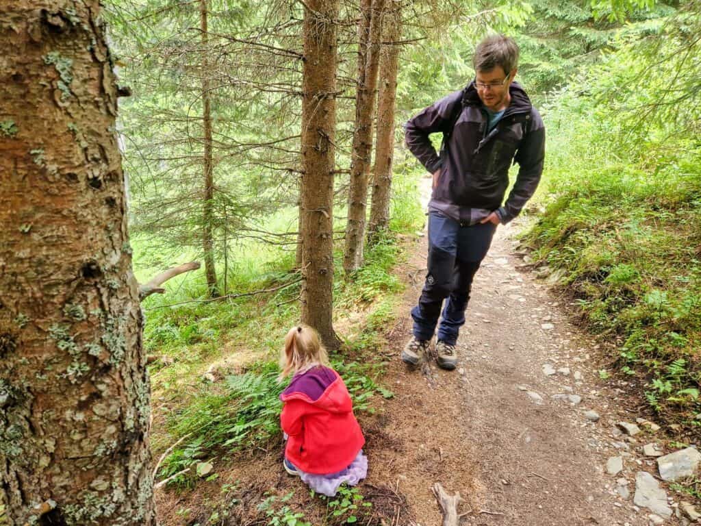 A man walks on a forest trail while a young child in a red jacket sits on the ground nearby, surrounded by green trees and foliage.