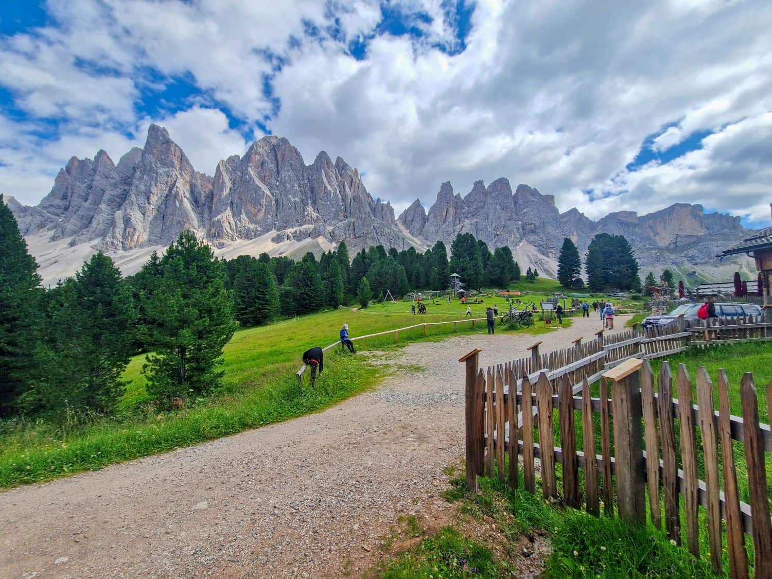 A scenic mountain landscape with jagged peaks, lush green grass, pine trees, a gravel path, and several people walking or relaxing outdoors under a partly cloudy sky. A wooden fence lines the right side of the image.