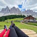 Two people lying on a wooden bench in a grassy field with a rustic cabin, pine trees, and dramatic mountain peaks in the background under a partly cloudy sky.