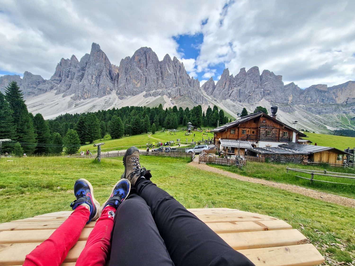 Two people lying on a wooden bench in a grassy field with a rustic cabin, pine trees, and dramatic mountain peaks in the background under a partly cloudy sky.