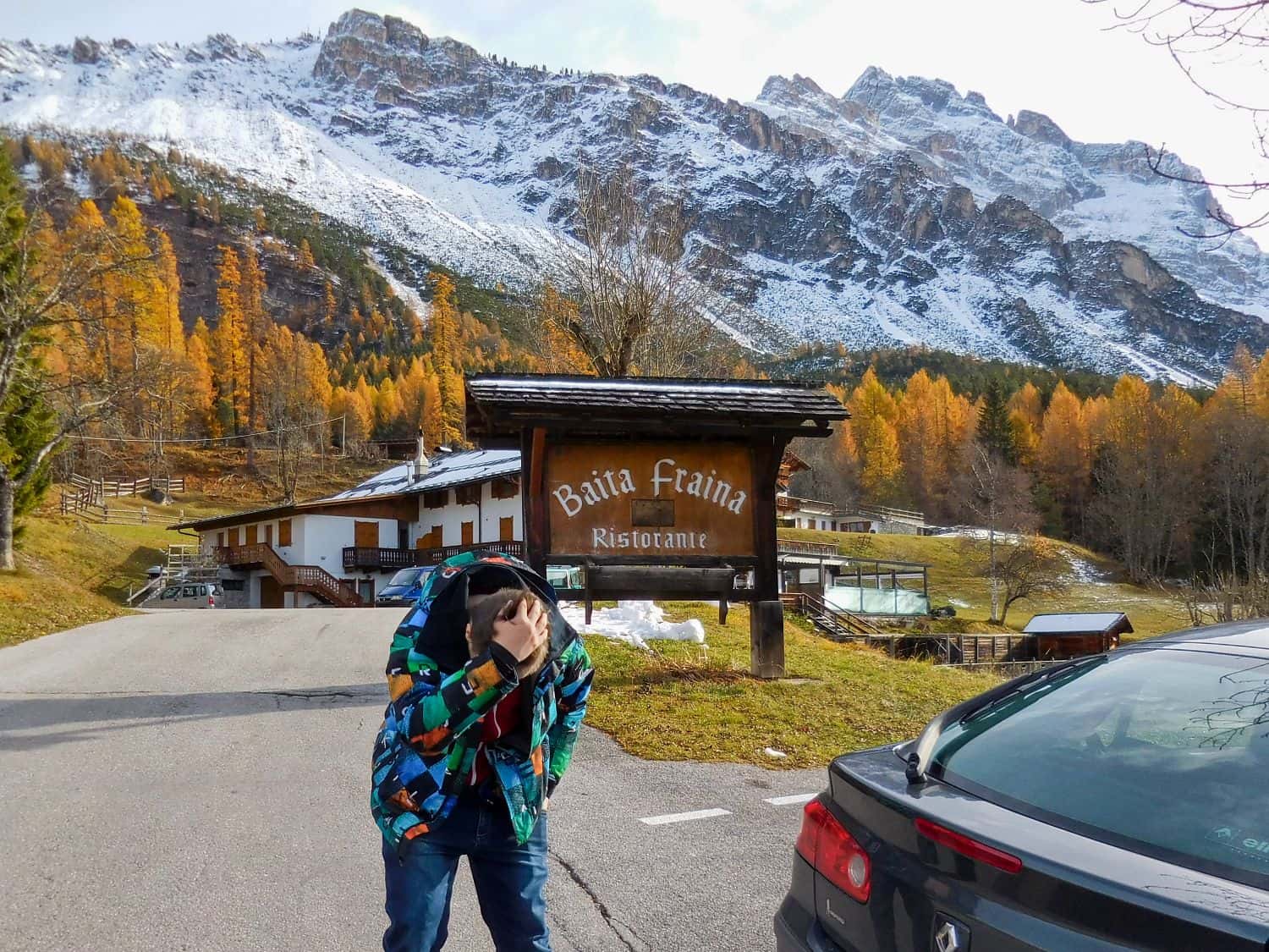 A person in a colorful jacket stands by a parked car, facing away and holding their head. Behind them is a wooden sign reading "Baita Fraina Ristorante," with snow-capped mountains and autumn trees in the background.