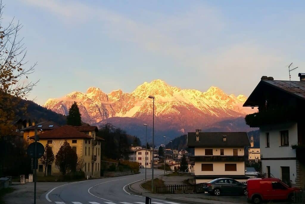 A curved road leads through a mountain village with houses in the foreground and snow-capped mountains in the background, illuminated by a warm, golden sunset. A red van is parked on the right side of the street.