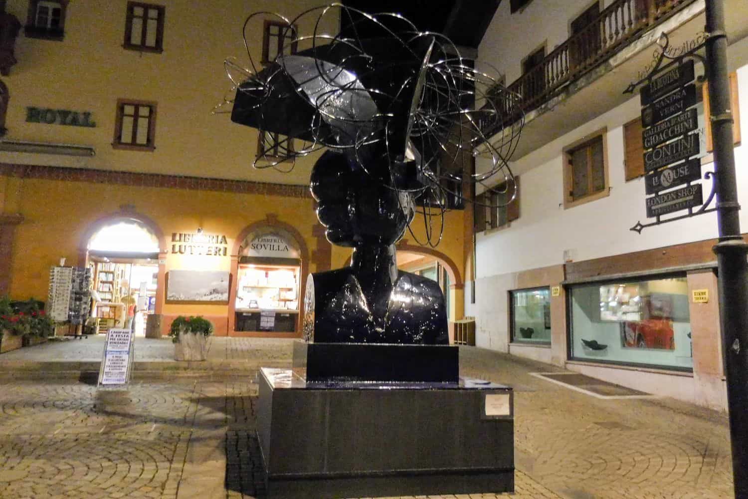 A shiny black sculpture of a human head with intricate metal wires swirling above it stands in a well-lit cobblestone square, surrounded by shops and buildings at night.
