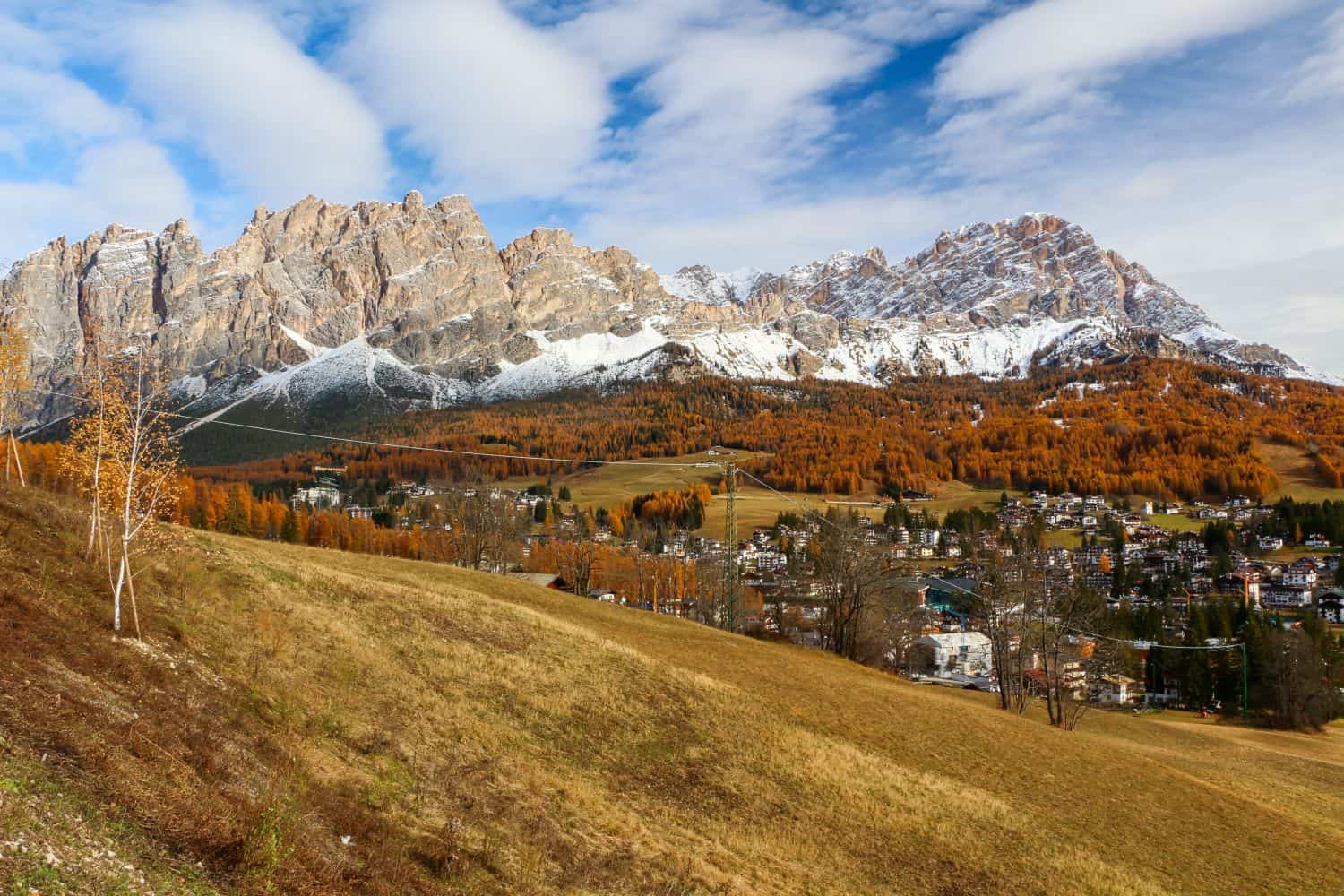 A panoramic view of a mountain range with rocky peaks, some dusted with snow, above a forest of autumn-colored trees and a small town nestled at the base under a partly cloudy sky.