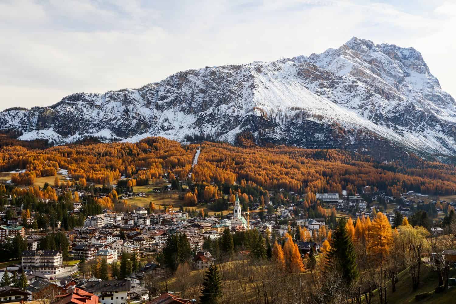 A picturesque mountain town sits below a large, snow-capped mountain; trees with golden autumn leaves cover the hills, and clusters of buildings fill the valley under a partly cloudy sky.