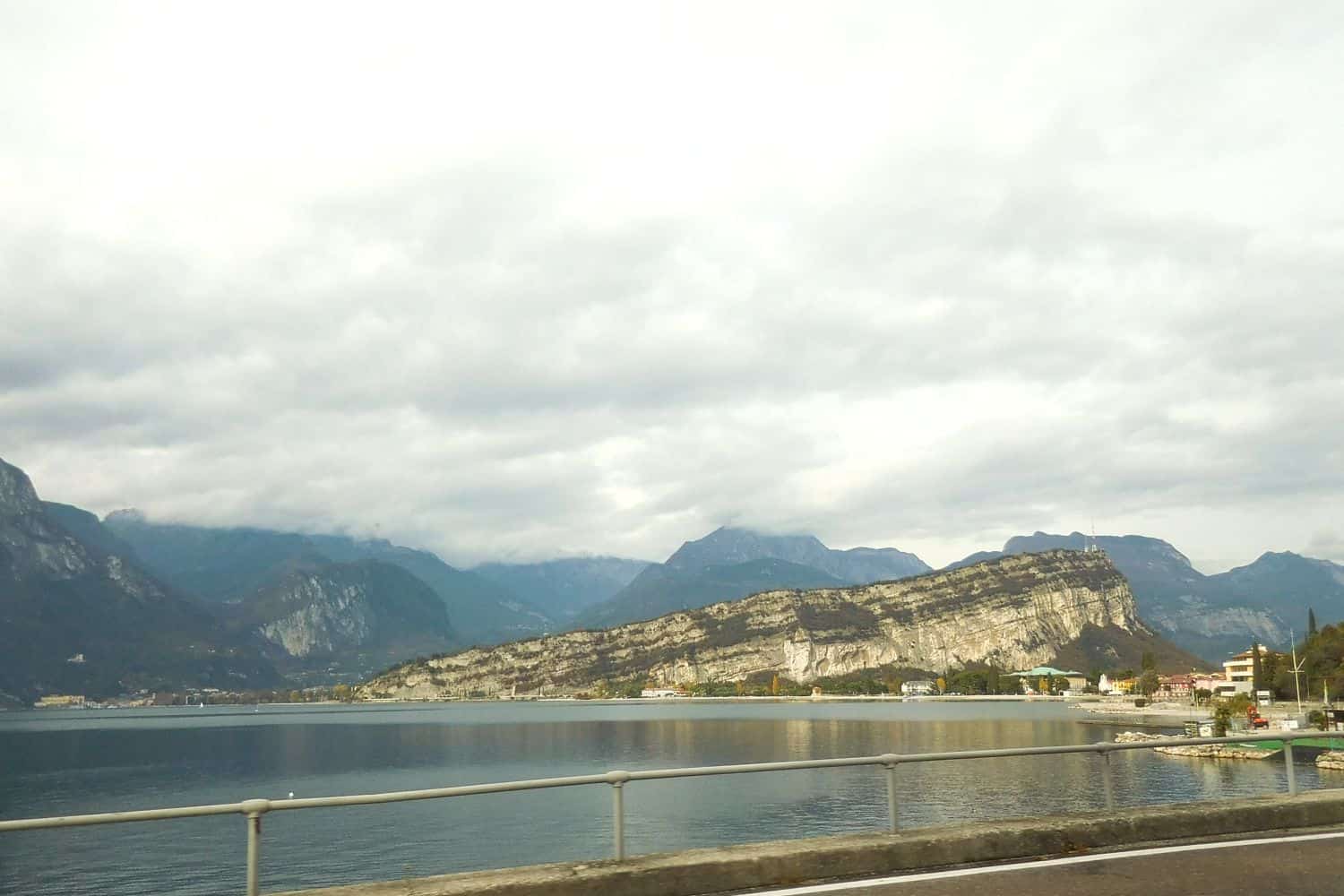 A scenic lakeside view with calm water, rugged mountains in the background, and cloudy skies above. A guardrail lines the road in the foreground.