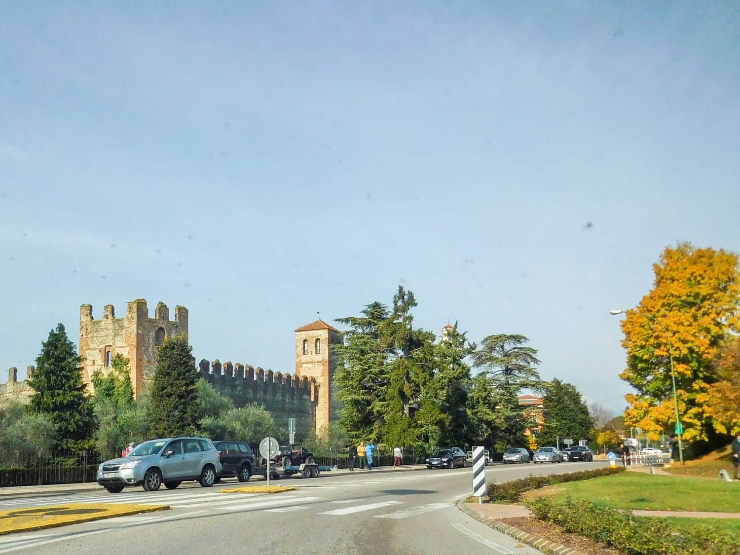 A castle-like stone wall with towers stands near a road roundabout lined with cars, trees, and people walking. The sky is clear, and autumn foliage is visible on the right side.