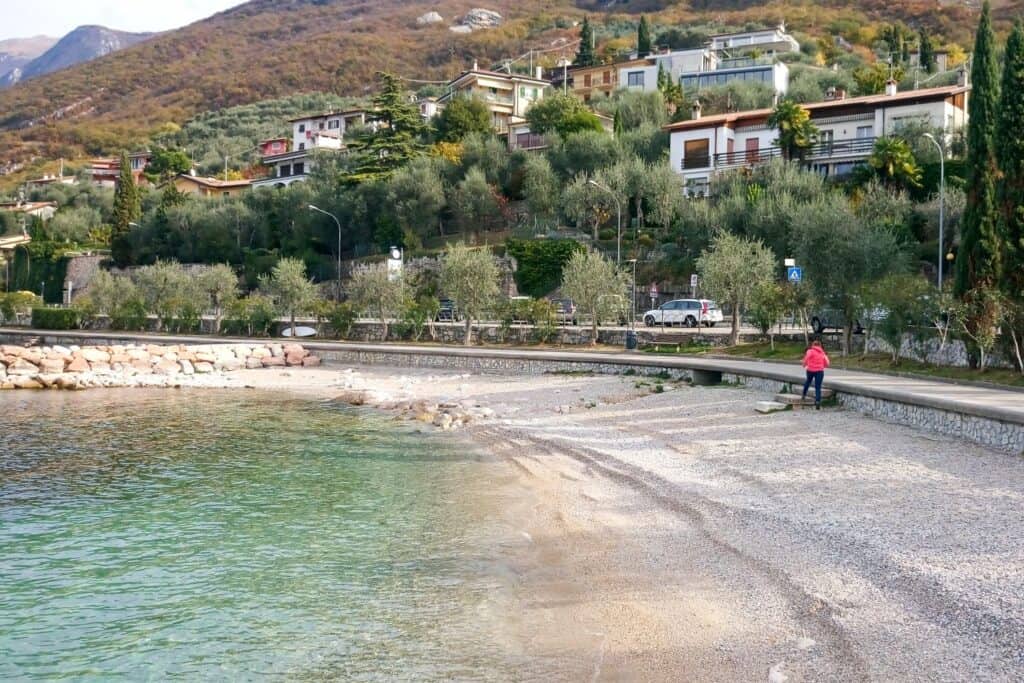 A person in a pink jacket sits on a stone wall by a pebbly beach with clear water. Houses and greenery cover the hillside in the background under a partly cloudy sky.