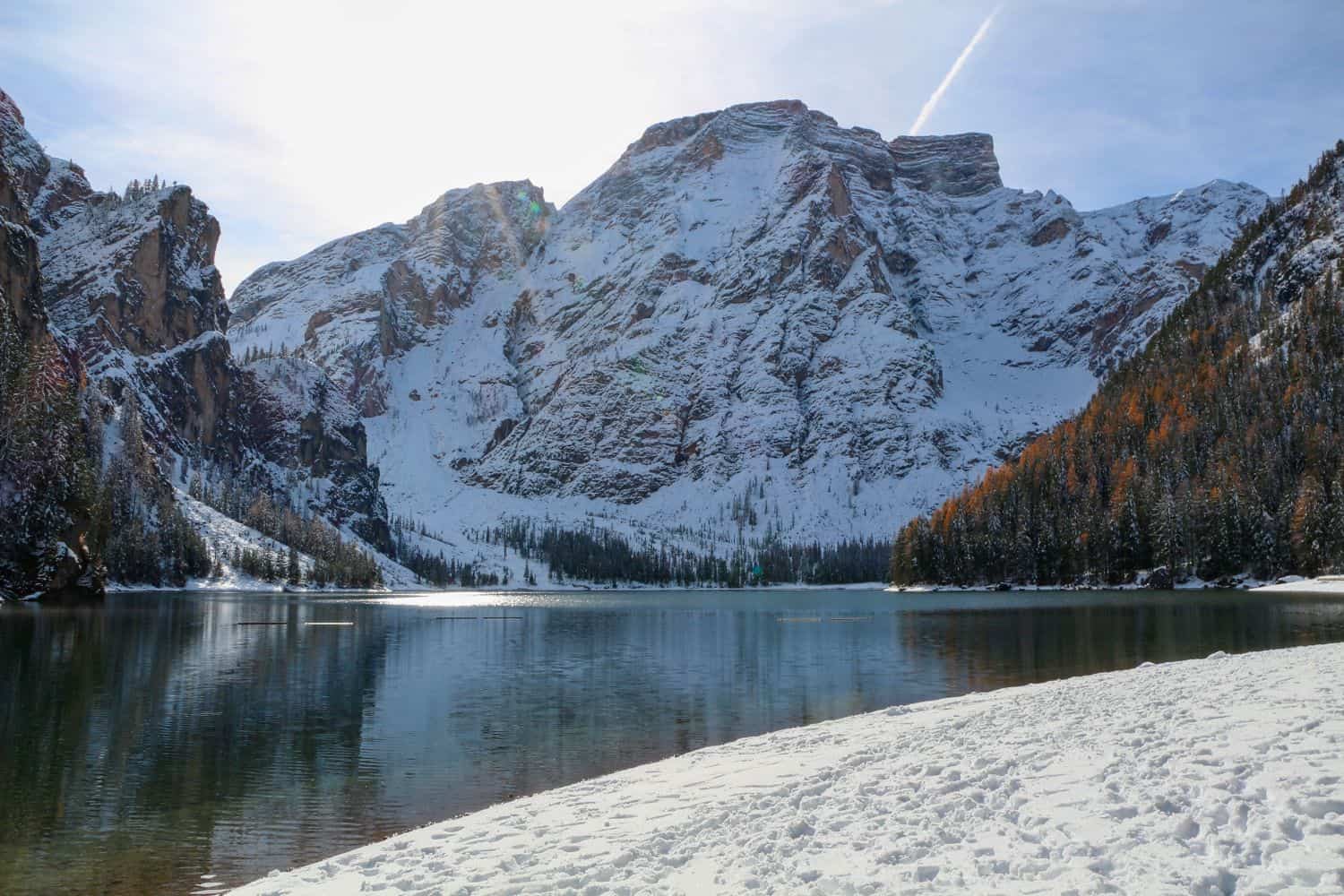 Snow-covered mountains rise above a calm lake, reflecting their snowy peaks. Pine trees with patches of autumn colors line the water’s edge, and the foreground shows snow on the shore under a bright, partly cloudy sky.