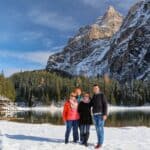 Four people bundled in winter clothes stand on snow by a lake, with trees and tall, snow-covered mountains in the background under a partly cloudy sky.