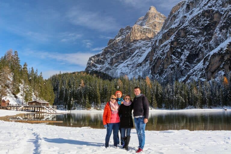 Four people bundled in winter clothes stand on snow by a lake, with trees and tall, snow-covered mountains in the background under a partly cloudy sky.