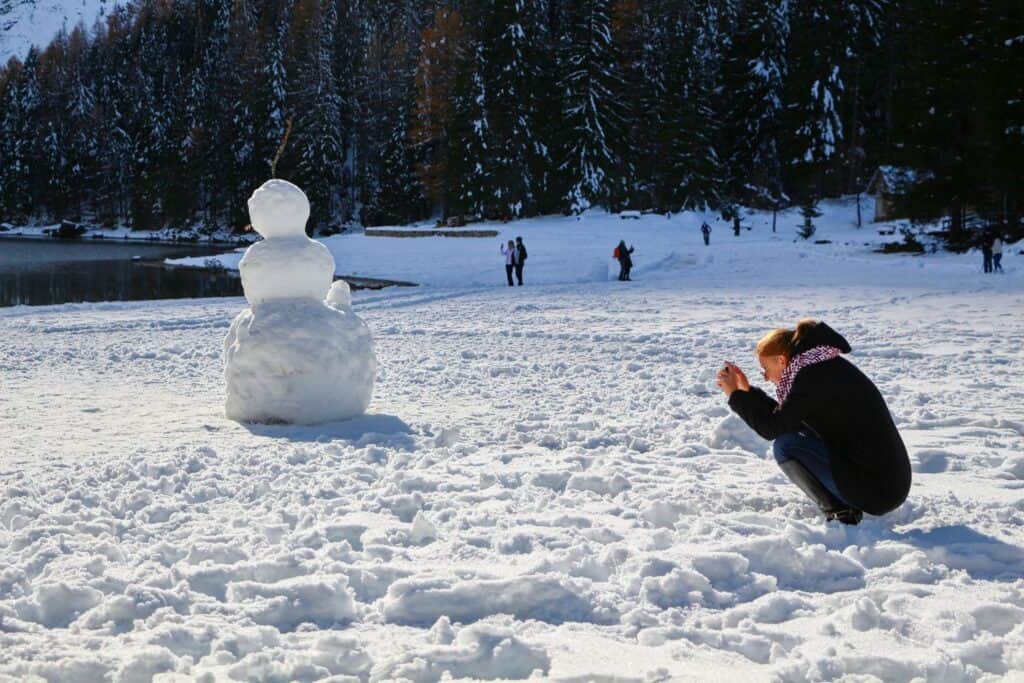 A person squats in the snow, taking a photo of a large snowman on a snowy field near a forested area, with other people in the background.