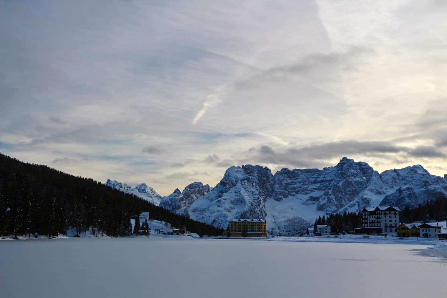 Snow-covered mountains under a cloudy sky, with buildings nestled at the base and a frozen lake in the foreground, surrounded by trees on one side.