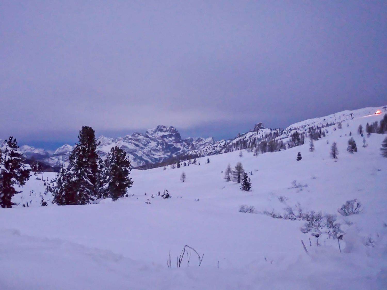 Snow-covered mountains and trees at dusk, with a pale blue and purple sky. A faint orange light glows on a distant slope, suggesting a cabin or chalet. The landscape is peaceful and serene.