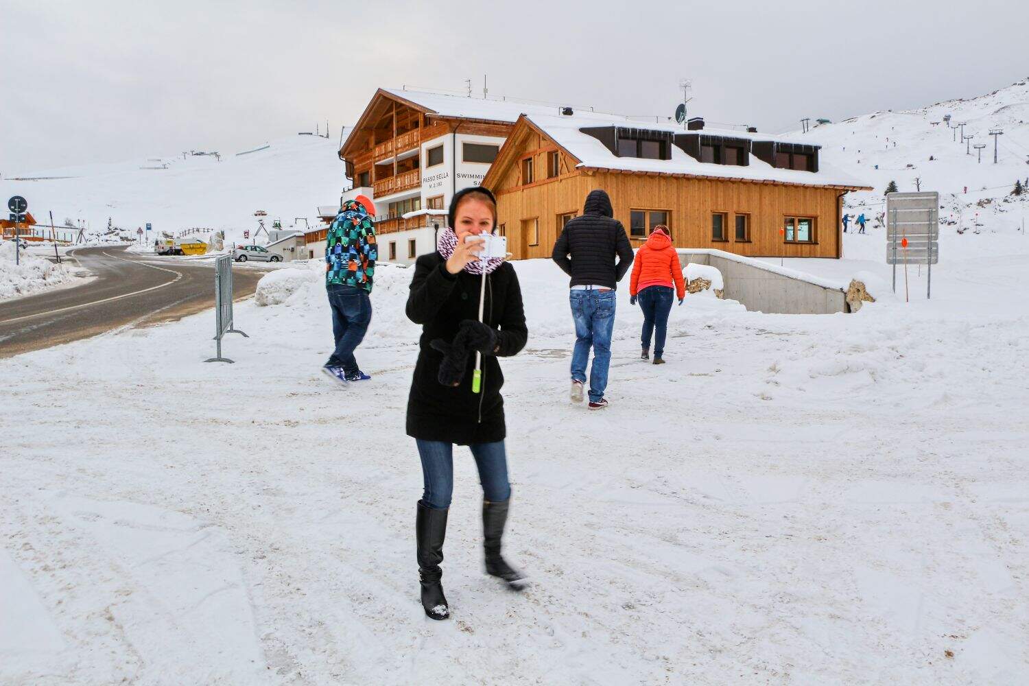 A person in a black coat and boots stands on snowy ground holding a striped lollipop, with a wooden building and several other people in winter clothing in the background.