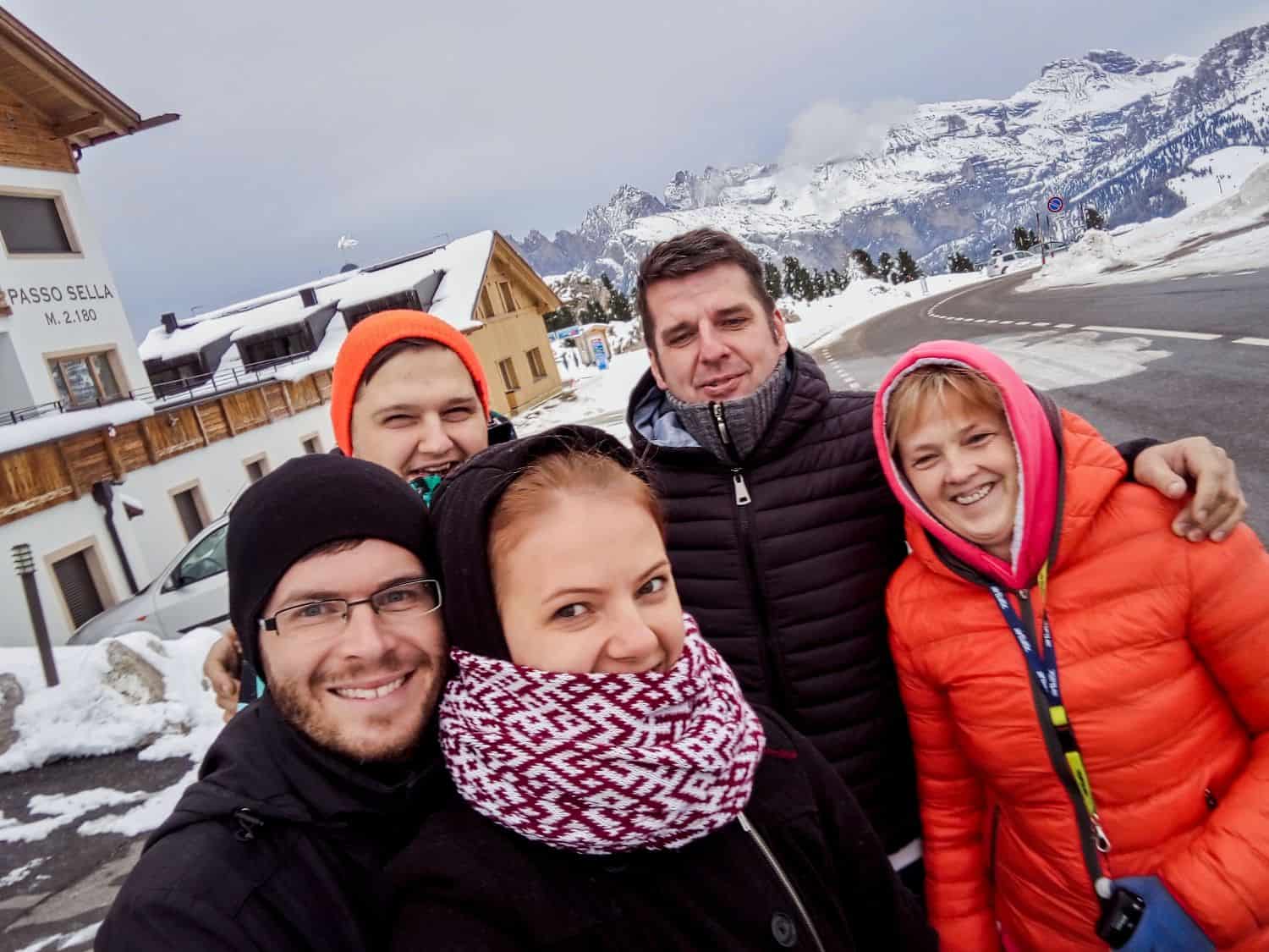 Five people bundled in winter clothing smile for a selfie with snowy mountains, a road, and wooden buildings in the background. The scene looks cold and cheerful.