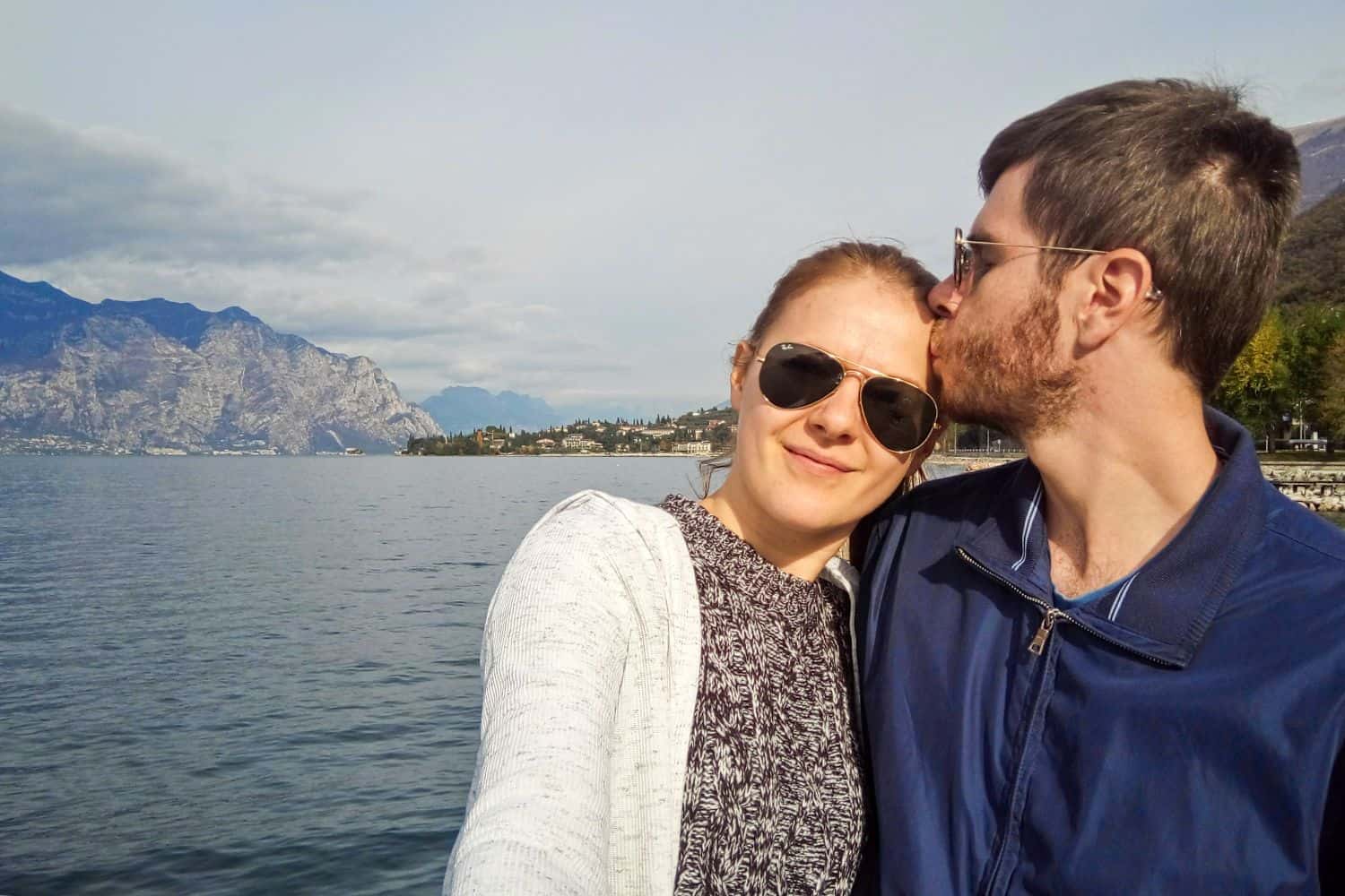 A couple wearing sunglasses takes a selfie by a lake, with mountains and a small town in the background. The man kisses the woman on the forehead as they both smile, enjoying a sunny day outdoors.