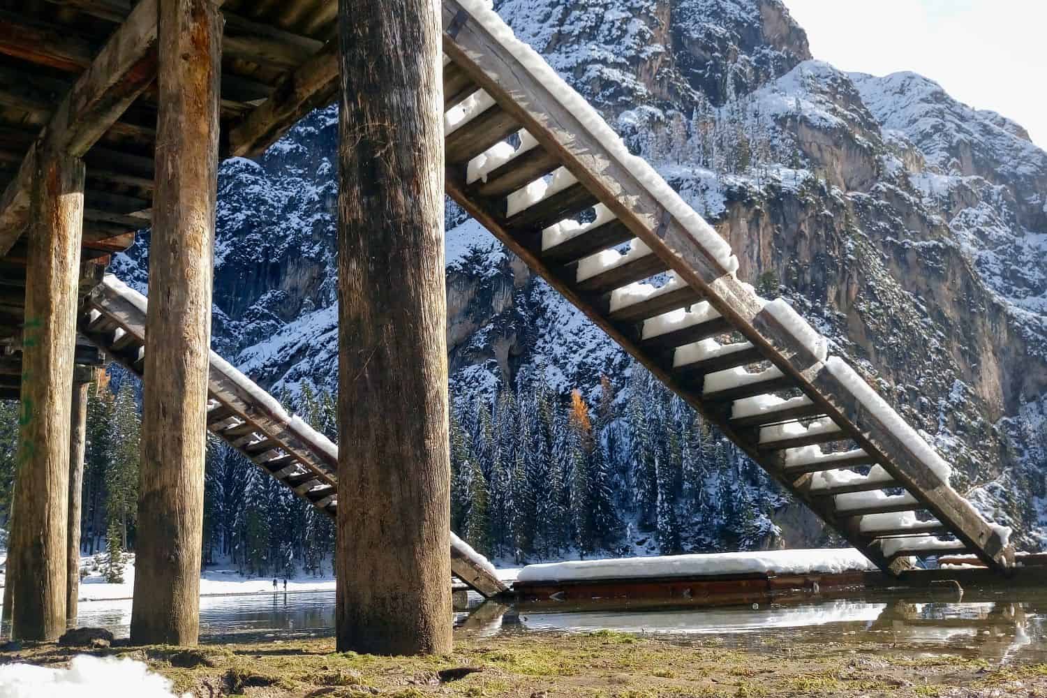 View from beneath a wooden staircase covered in snow, with tall support beams, snowy mountains, and pine trees in the background against a partly cloudy sky. The ground below is wet and partially frozen.