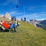 A person relaxes on a lounge chair on a grassy mountain slope, while others walk nearby. A flag waves on a tall pole. Rocky mountains and a blue sky are in the background.