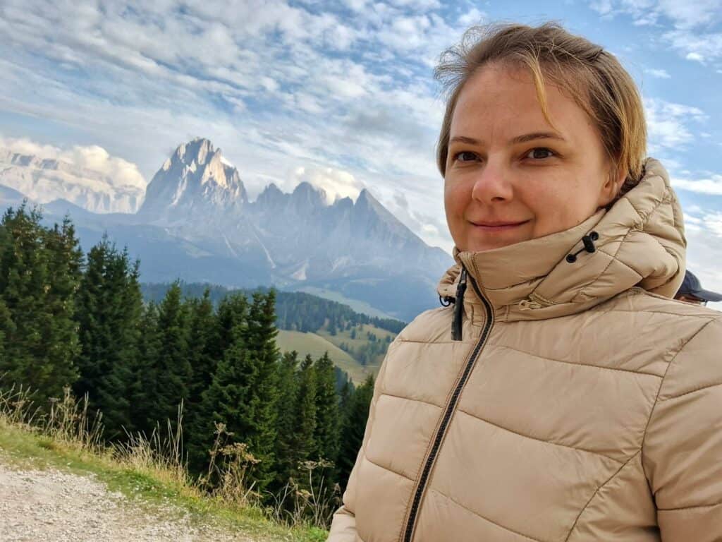 A woman in a beige puffer jacket stands outdoors, smiling slightly, with green pine trees and rugged, snow-dusted mountains in the background under a partly cloudy sky.