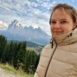 A woman in a beige puffer jacket stands outdoors, smiling slightly, with green pine trees and rugged, snow-dusted mountains in the background under a partly cloudy sky.