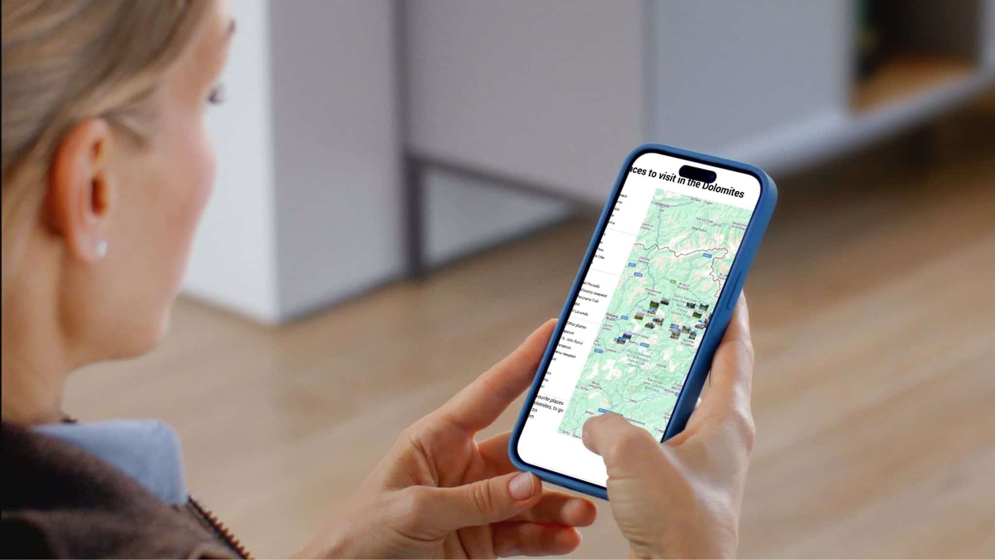 A person holds a smartphone displaying a map application with various locations marked, while seated indoors on a wooden floor.