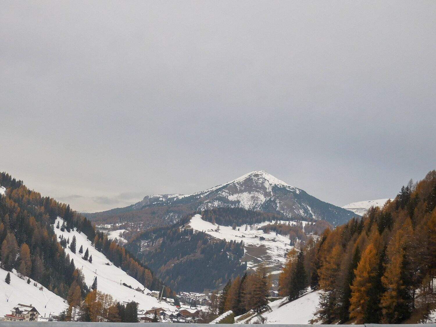 A snow-covered mountain rises in the distance, surrounded by evergreen and autumn-colored trees. Small houses are scattered along the snowy hillsides under a cloudy gray sky.