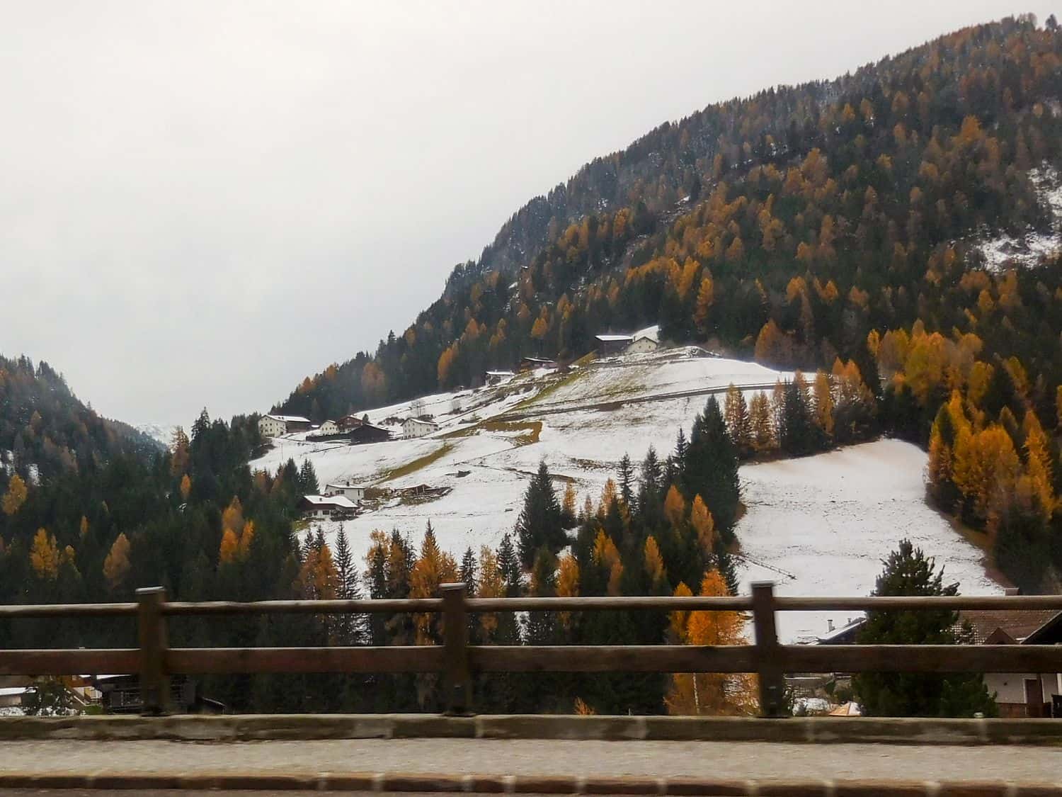 A hillside with scattered houses and snow-covered fields, surrounded by autumn-colored trees and forested mountains, seen from behind a wooden fence or railing. The sky is overcast.