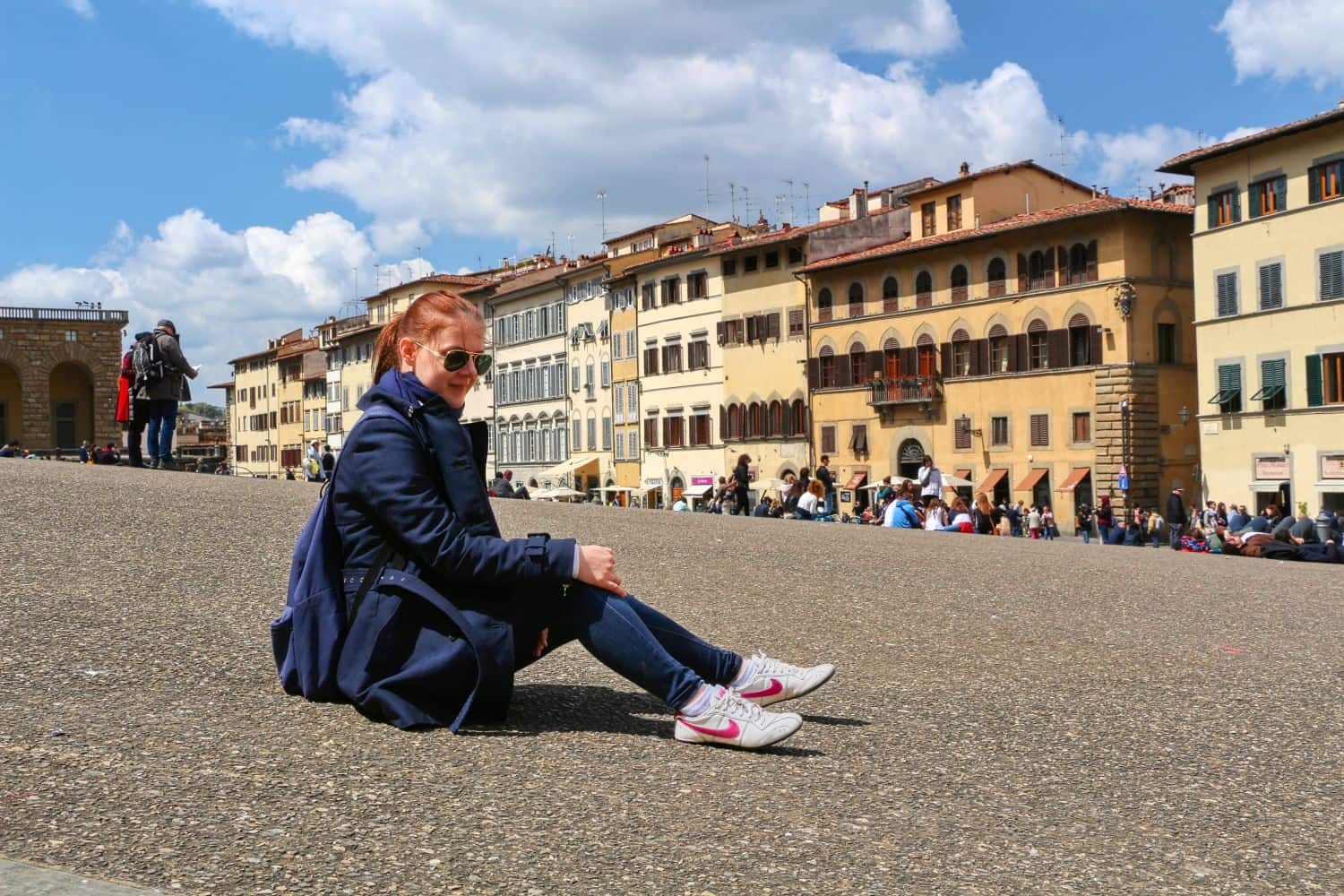 A person in a navy coat, jeans, and white sneakers sits on a stone plaza with historic yellow buildings and many people in the background under a partly cloudy sky.