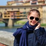 A woman wearing sunglasses, a navy coat, and a scarf stands by a stone railing with a blurred view of the Ponte Vecchio bridge in Florence, Italy, and a river in the background.