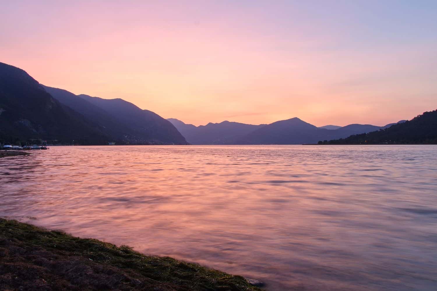 A calm lake at sunrise with gentle waves, distant mountains silhouetted against a pink and orange sky, and a grassy shoreline in the foreground.