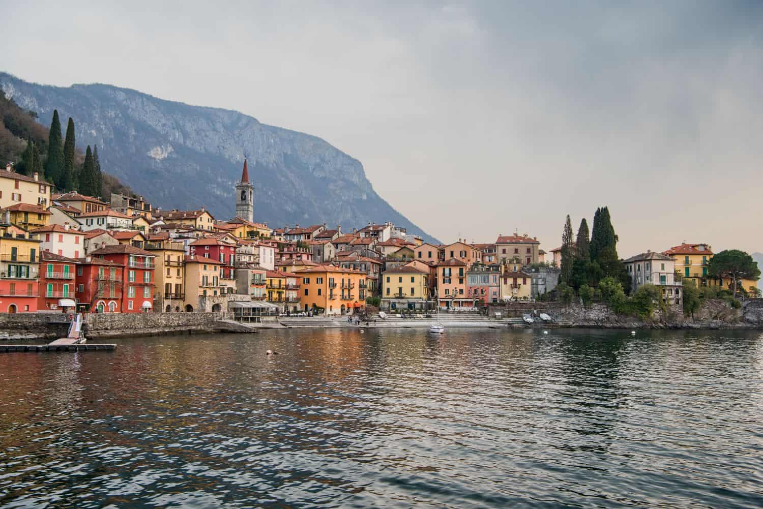 A picturesque lakeside village with colorful buildings, a church tower, and cypress trees, set against mountains and reflected in calm water under a cloudy sky.