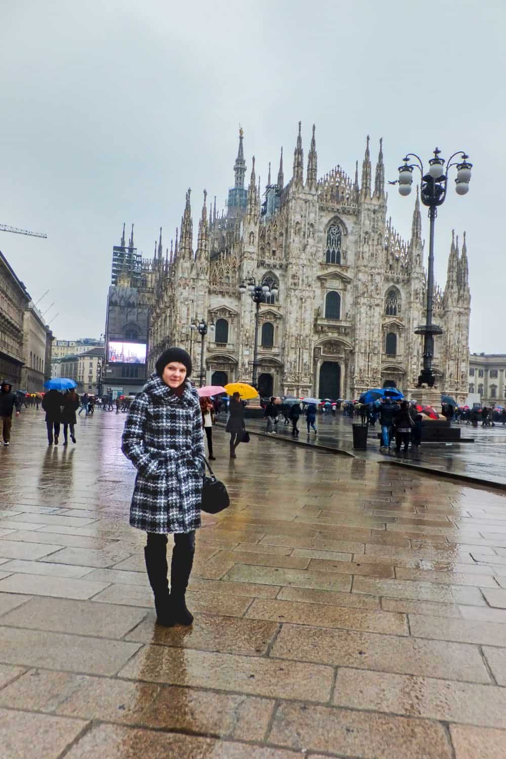 A woman in a plaid coat and black boots stands smiling on a wet plaza in front of Milan Cathedral (Duomo di Milano) on a rainy day, with people holding colorful umbrellas in the background.