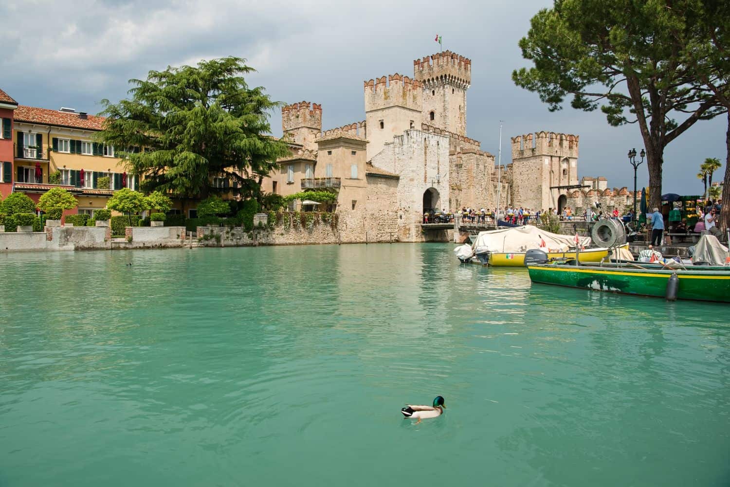 A duck swims in teal water near colorful buildings and a historic stone castle with towers and battlements. Boats are docked nearby, and trees and people line the waterfront under a cloudy sky.