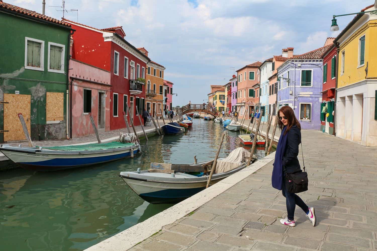 A woman walks along a canal lined with colorful buildings and boats in Burano, Italy, on a cloudy day. The houses are painted in vibrant red, yellow, purple, and green.