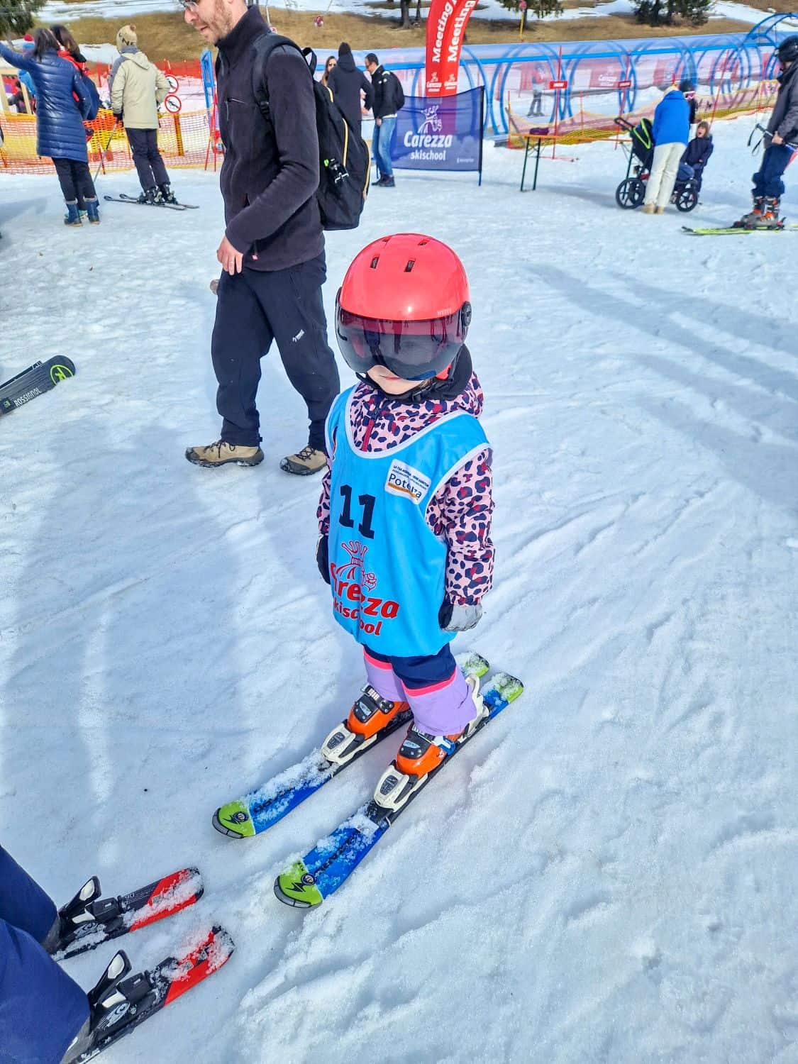 A young child in a red helmet, blue ski vest, and colorful jacket stands on skis in the snow, ready for a ski lesson. Adults and other skiers are nearby on a snowy slope with equipment and banners visible in the background.