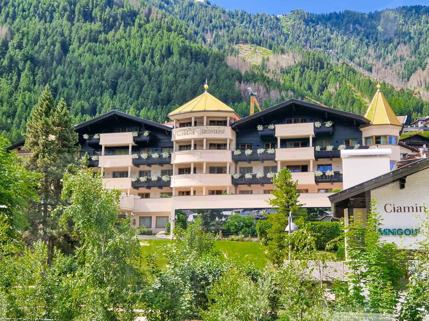 A large alpine hotel with balconies and gold-tipped turrets sits among green trees, set against a forested mountain backdrop under a clear blue sky.