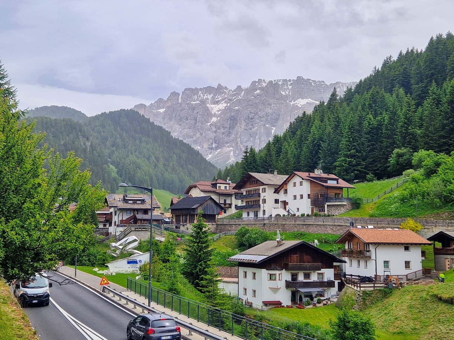A scenic mountain village with alpine houses, pine trees, and a winding road with cars. Snow-capped peaks rise in the background under a cloudy sky.