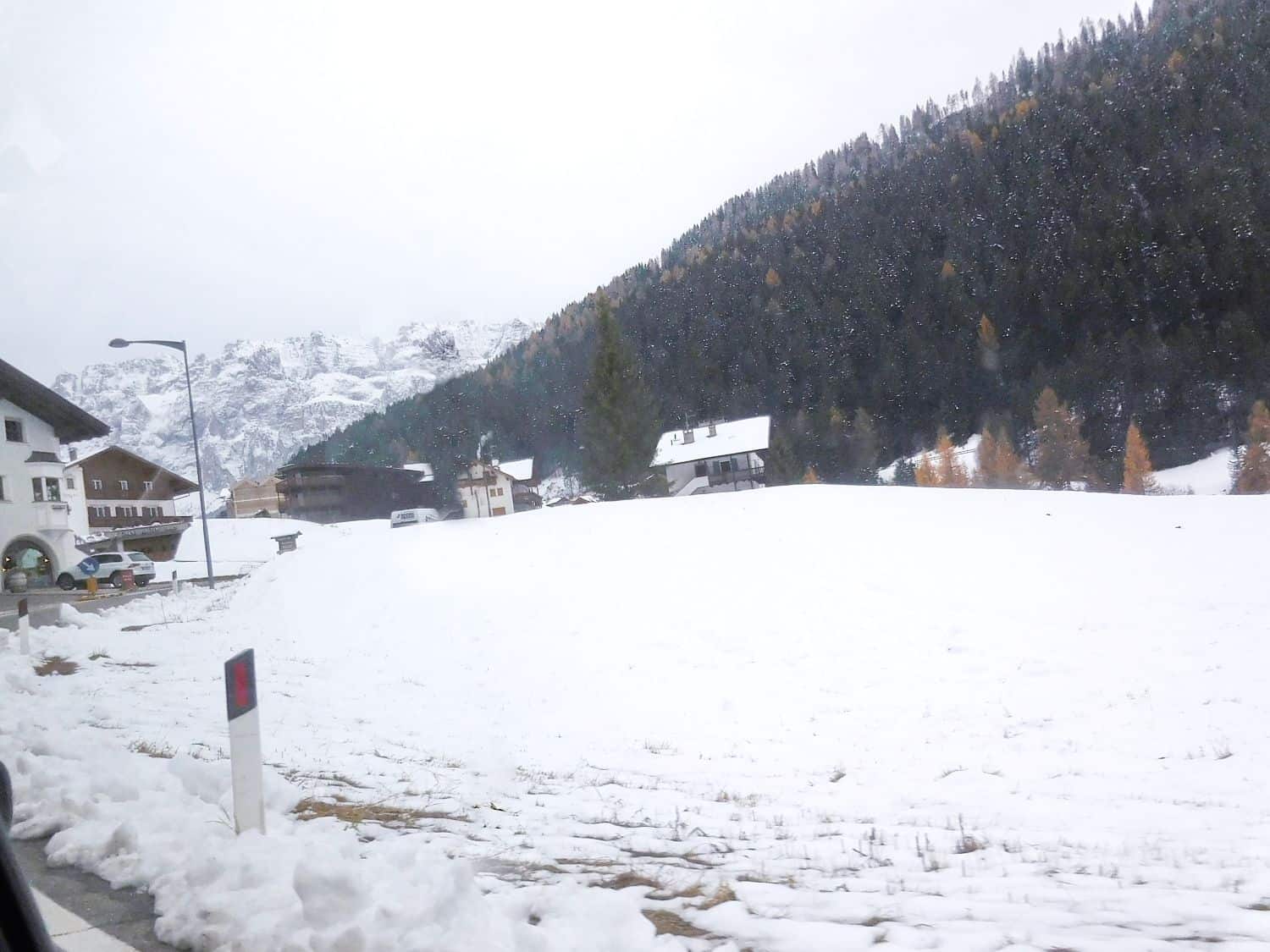 Snow-covered landscape with a few houses scattered near the base of a forested mountain. Evergreen trees cover the slope, and some buildings are partly visible on the left. The sky appears overcast.